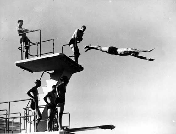 photo of man in mid-air after diving off a high dive board, while others watch from the steps behind him.
